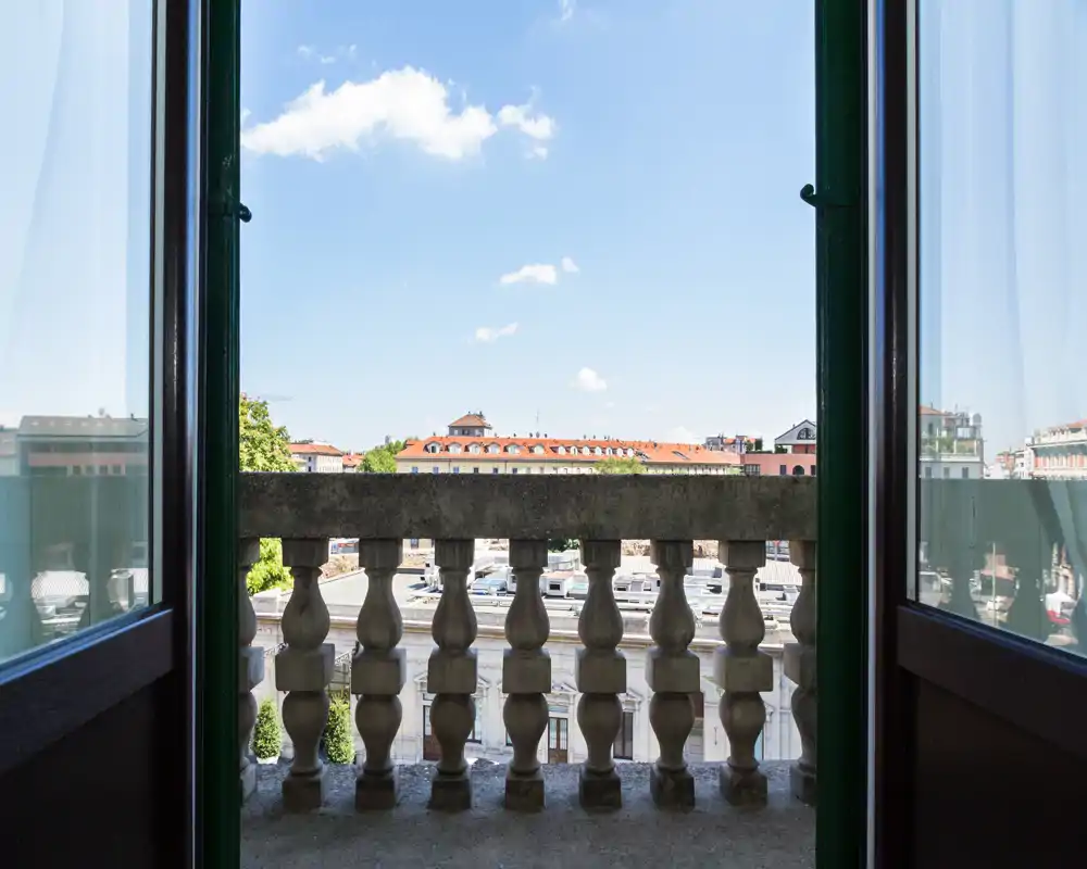 Sunny balcony view framed by an open double door and classic stone balustrade, overlooking city rooftops and blue sky — ideal for morning coffee.