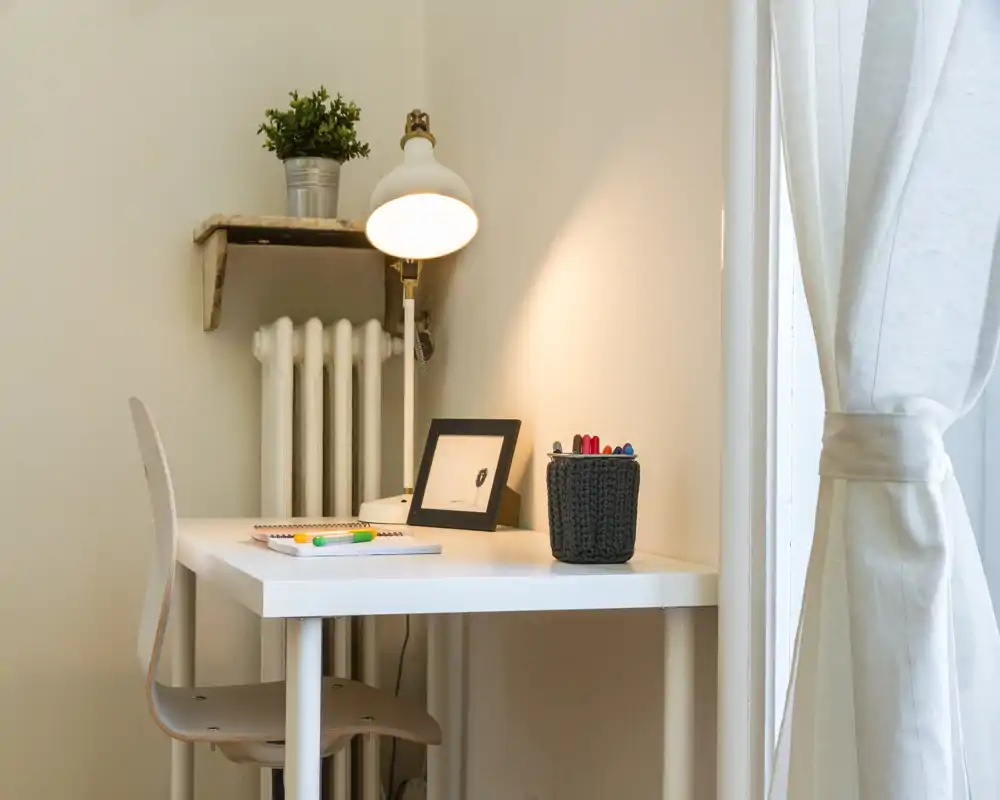 Small, tidy workspace corner with a white desk, chair, desk lamp and natural light from the window — ideal for remote work or study.