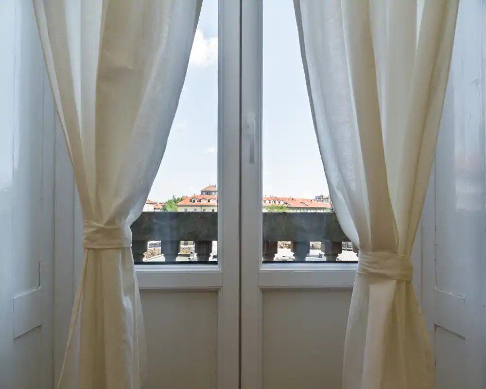 View through a glass door framed by light curtains onto a small balcony and distant rooftops — bright and airy city outlook.
