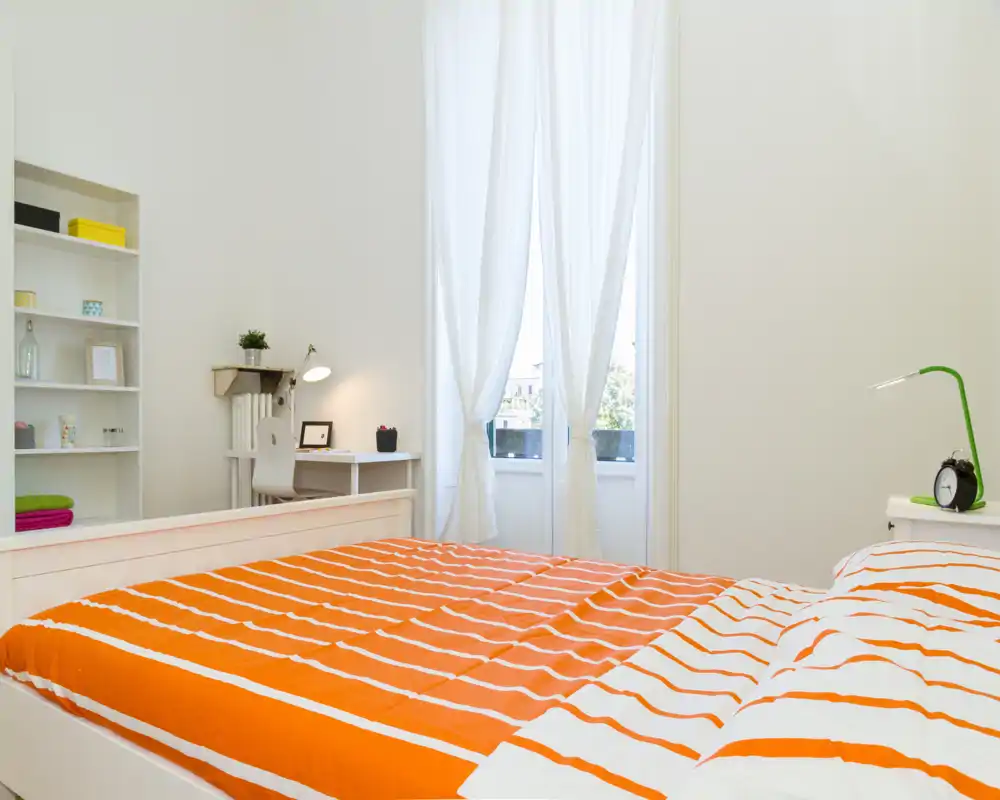Bright, modern bedroom with a large bed dressed in orange-striped bedding, a bedside table with a lamp and clock, and a work desk near a tall window with sheer curtains letting in natural light.