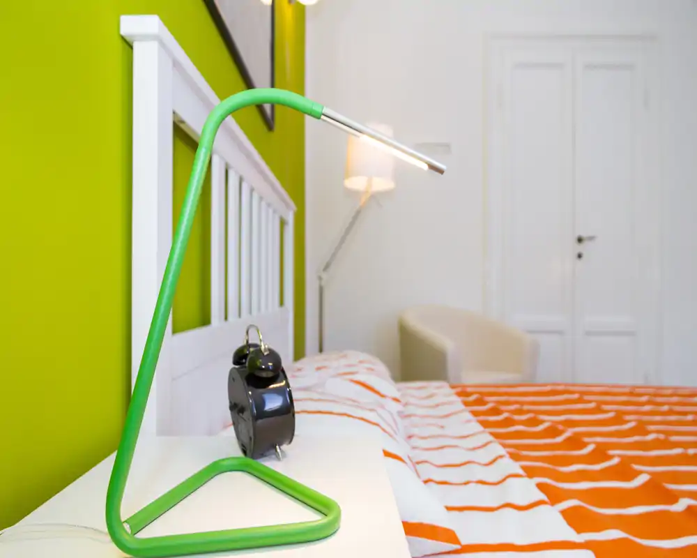 Bright, modern bedroom with a white headboard, orange patterned bed linen and a green bedside lamp creating a fresh, colorful look.