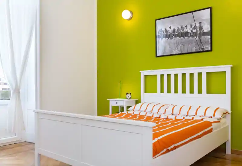 Bright, modern bedroom with a white wooden bed against a vivid green accent wall, crisp orange-striped bedding and natural light from a tall window.
