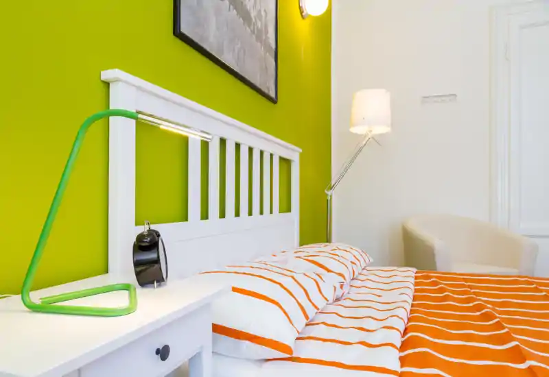 Bright, modern bedroom with a white slatted headboard, orange-striped bedding and a green bedside lamp; cozy, well-lit and stylish.