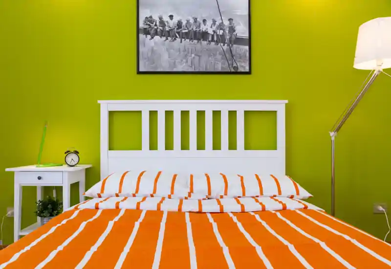 Bright, modern bedroom with a white slatted headboard and an orange-and-white striped duvet. The room features a bedside table with alarm clock and a tall floor lamp, set against a vivid green accent wall.