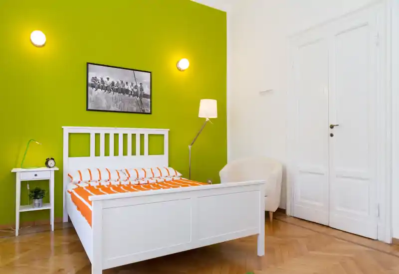 Bright, modern bedroom with a white wooden bed against a vivid green accent wall, bedside table, floor lamp and a cozy armchair; clean parquet floor and simple, contemporary styling.