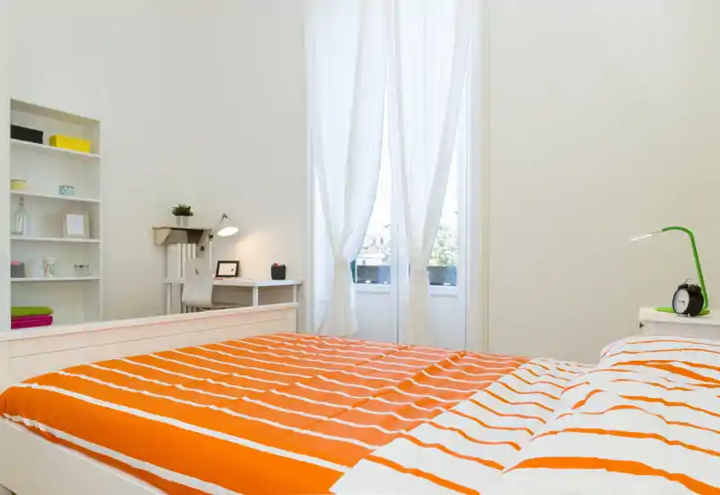 Bright, modern bedroom with a large bed dressed in orange-striped bedding, a bedside table with a lamp and clock, and a work desk near a tall window with sheer curtains letting in natural light.