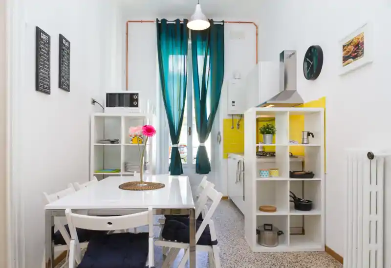 Sunny dining area integrated with a compact kitchen: white table with folding chairs, decorative flowers and cheerful yellow backsplash behind the hob. Ideal for small groups and cozy meals with natural light from the balcony door.