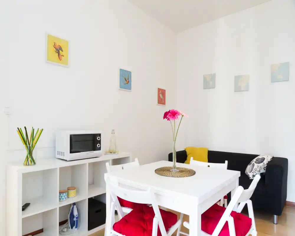 Rincón salón/comedor luminoso y moderno con mesa blanca, sillas plegables con cojines rojos, sofá negro pequeño y cuadros alegres — perfecto para un alquiler compacto y con estilo.