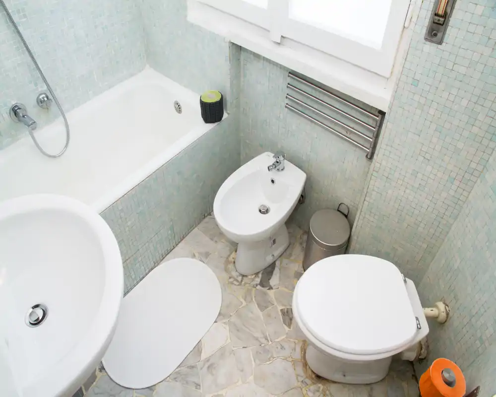 Baño compacto y limpio con bañera, inodoro y bidé; azulejos en mosaico claros y suelo tipo mármol que aportan luminosidad.