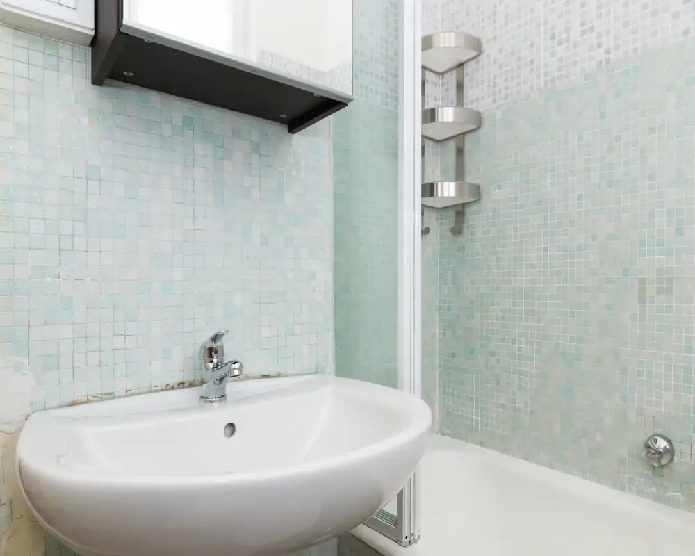 Baño compacto con lavabo cerámico blanco, grifo cromado y área de bañera/ducha con mosaico de azulejos azul claro. Espacio limpio y de estilo sencillo con mampara de vidrio.