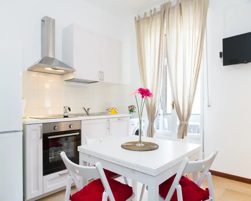 Cocina compacta y luminosa con mobiliario blanco, horno empotrado y una pequeña mesa con cojines coloridos y flores frescas — ideal para un apartamento urbano acogedor.