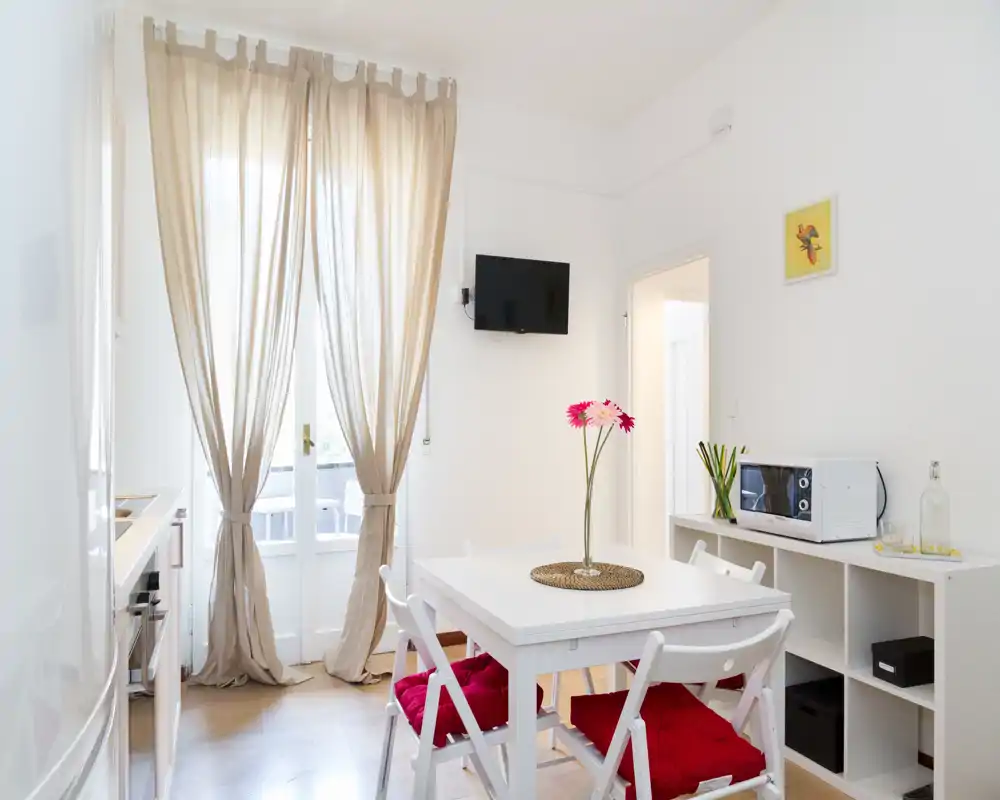 Área de comedor luminosa y acogedora con mesa blanca, sillas con cojines rojos y un jarrón de flores junto a la puerta del balcón iluminada.