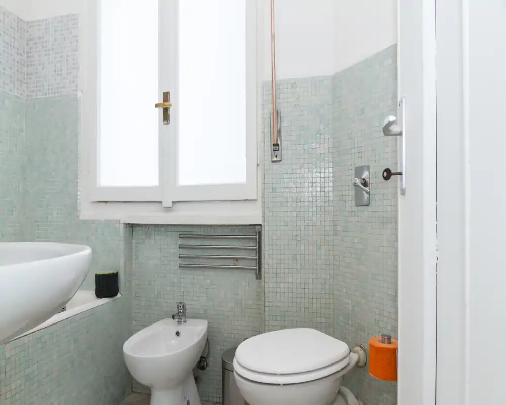 Baño compacto alicatado con inodoro, bidé y lavabo bajo una ventana con vidrio esmerilado; mosaico aqua claro y grifería cromada para un espacio funcional y limpio.