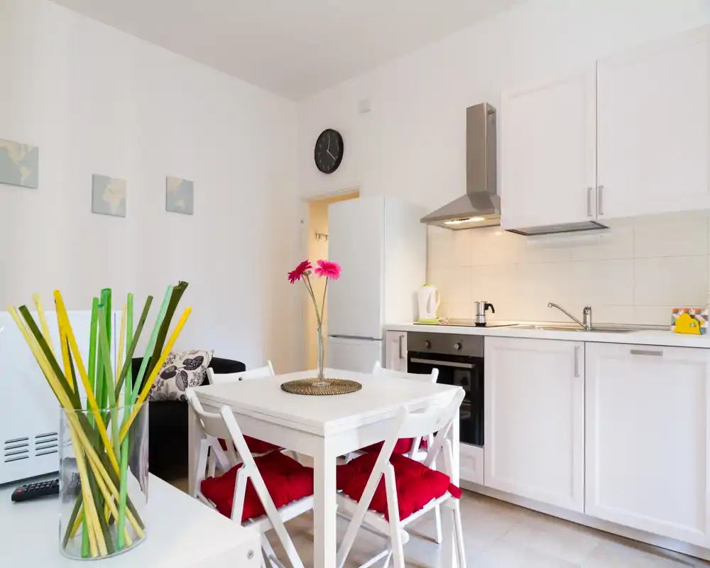 Cocina abierta moderna y luminosa con muebles blancos, horno integrado, frigorífico y una mesa pequeña con cojines rojos — bien decorada y acogedora.