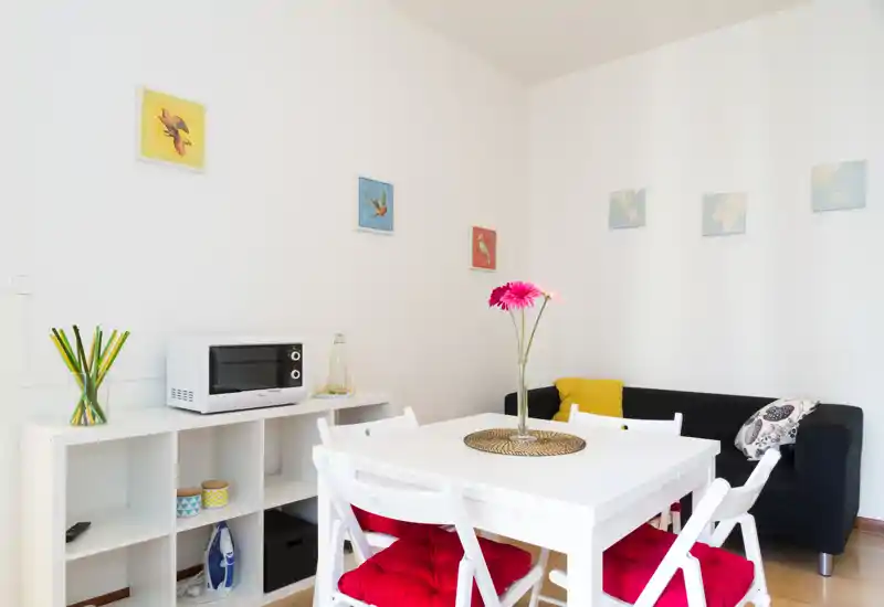 Rincón salón/comedor luminoso y moderno con mesa blanca, sillas plegables con cojines rojos, sofá negro pequeño y cuadros alegres — perfecto para un alquiler compacto y con estilo.