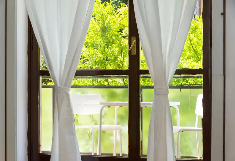 Bright balcony view framed by white curtains, with outdoor chairs and lush green foliage visible through the glass door.