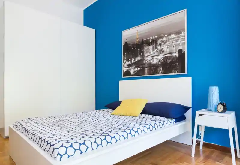 Bright, modern bedroom with a white platform bed against a vivid blue accent wall, patterned bedding, bedside table with lamp and alarm clock — clean, stylish and well lit.