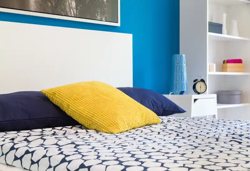 Bright, modern bedroom with patterned bedding, a prominent yellow cushion and a clean white headboard; neatly styled shelving and bedside table create a fresh, inviting atmosphere.