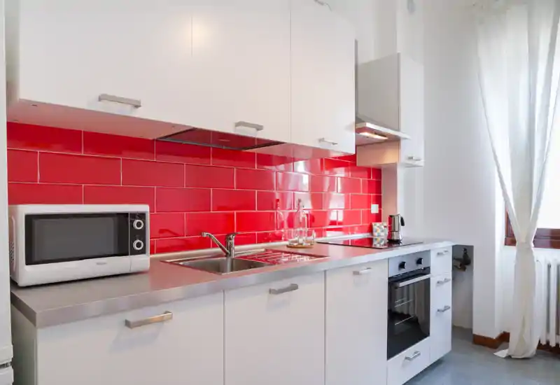 Cuisine moderne et compacte avec crédence en carrelage rouge vif, plan de travail en inox, four encastré, évier et micro-ondes. La lumière naturelle venant de la fenêtre rend l'espace lumineux et accueillant.