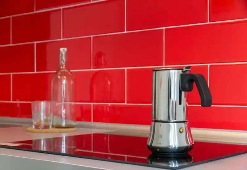 Plan de travail de cuisine moderne et lumineux avec plaque de cuisson et cafetière moka en inox devant des carreaux rouges — propre, élégant et prêt pour le café.