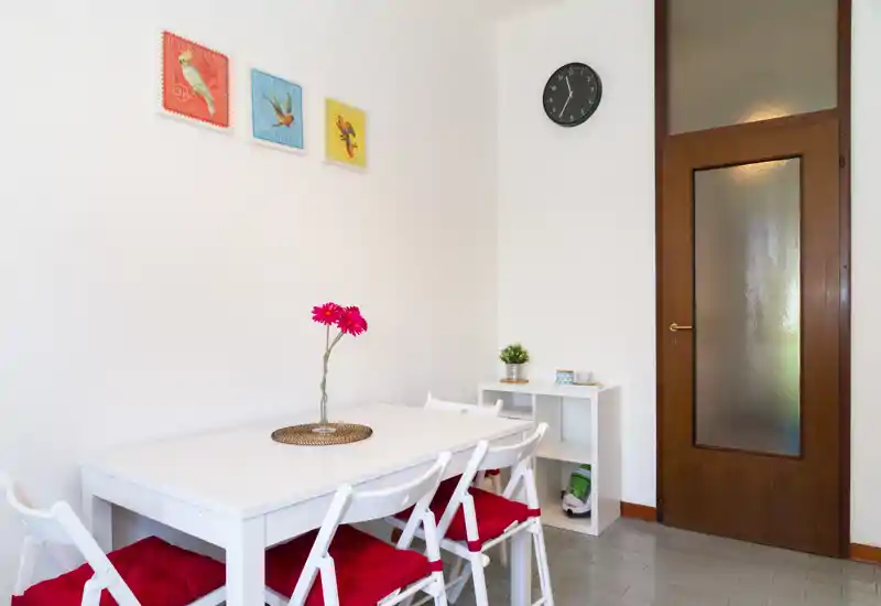 Coin repas lumineux et épuré avec table blanche, quatre chaises aux coussins rouges et un centre de table floral, complété par des tableaux muraux et une petite étagère.