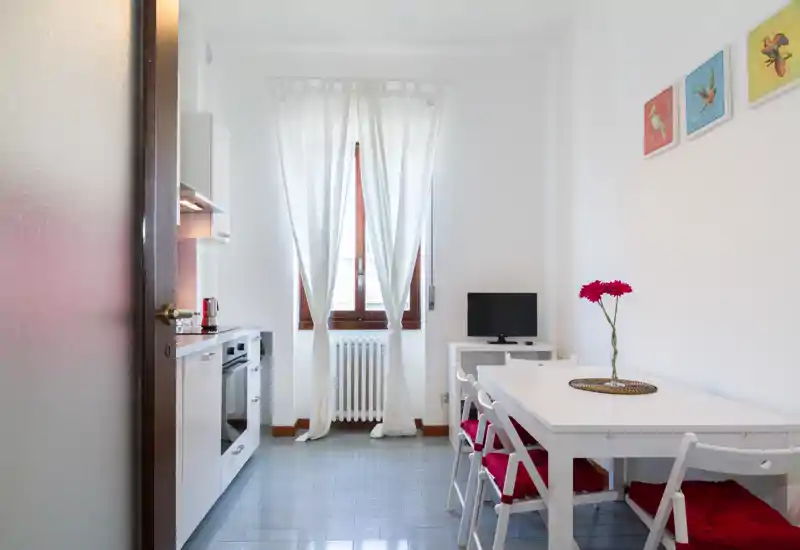 Coin cuisine-salle à manger compact et moderne avec des meubles blancs à gauche, une table avec chaises à coussins rouges à droite et une fenêtre lumineuse au fond. Espace propre et bien éclairé, idéal pour couples ou petits groupes.