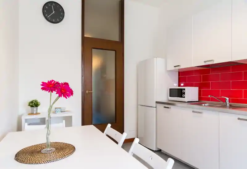 Kitchenette moderne et lumineuse avec placards blancs, crédence rouge carrelée, réfrigérateur, micro-ondes et table décorée de fleurs fraîches — idéale pour les petits appartements urbains.