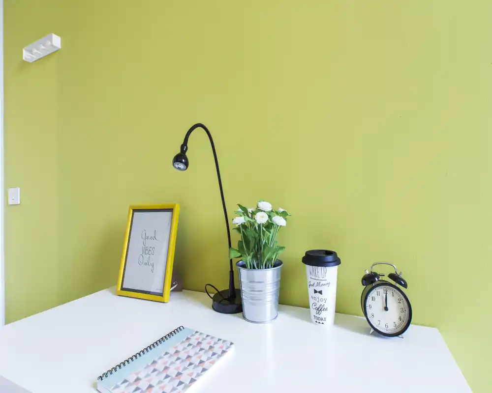 Rincón de escritorio luminoso y minimalista con mesa blanca frente a una pared verde, decorado con lámpara, marco, planta en maceta, vaso para llevar y despertador — imagen de un espacio de trabajo ordenado.