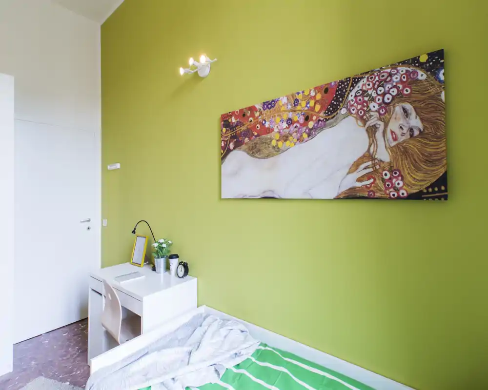 Rincón de dormitorio luminoso y moderno con cama individual, escritorio blanco y arte colorido en una pared verde; ordenado y acogedor para estancias cortas.
