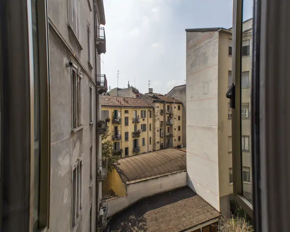 Vista de un patio interior y edificios residenciales circundantes desde una ventana abierta, con fachadas cálidas y detalles de tejados.