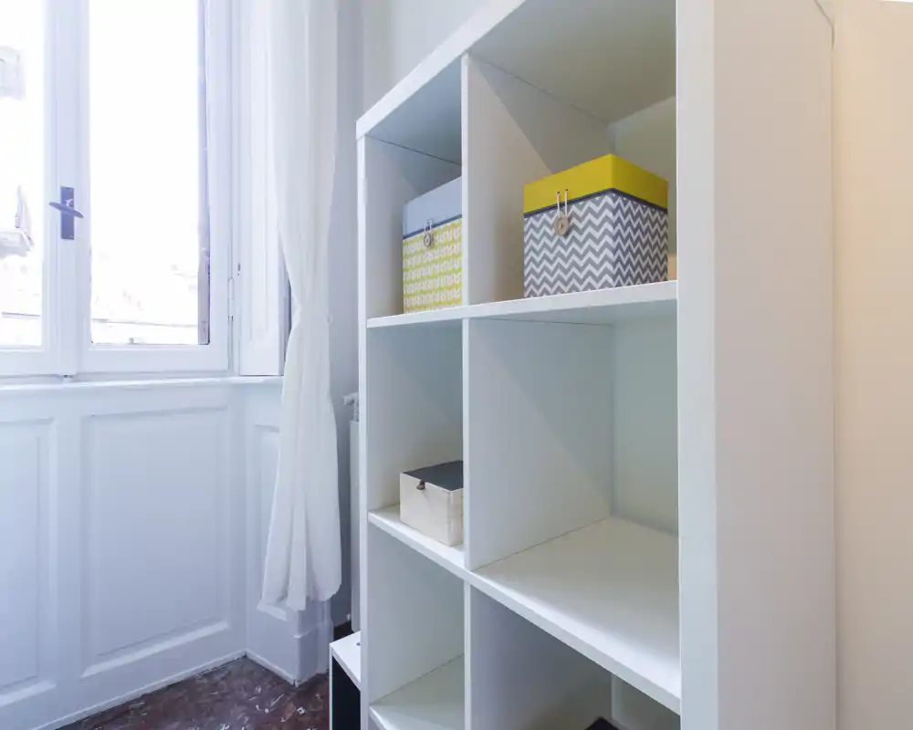 Alcoba de entrada luminosa con estanterías blancas y una ventana alta que aporta luz natural; cajas de almacenamiento que mantienen el espacio ordenado y funcional.