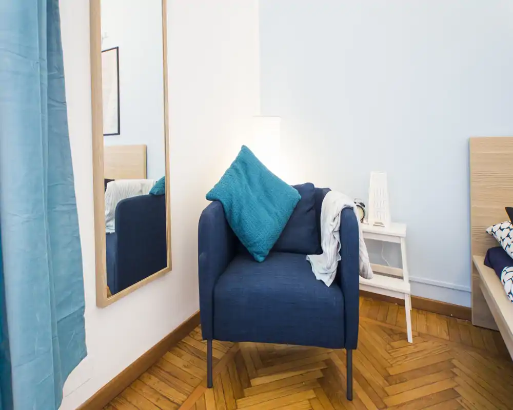 Coin chambre lumineux et moderne avec un fauteuil bleu, coussin turquoise et une table de chevet ; tête de lit partielle et miroir mural visibles.