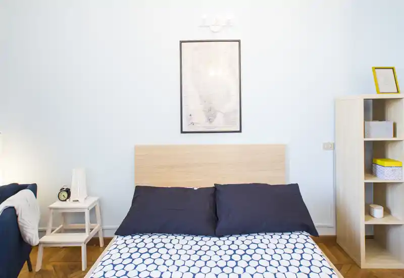 Camera da letto luminosa e dal design essenziale con un letto matrimoniale, piumone a fantasia e cuscini blu. Testiera e scaffale in legno chiaro per un aspetto pulito e minimale.