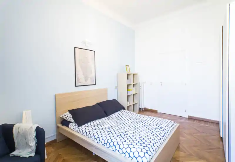 Camera da letto luminosa e minimal con letto matrimoniale, copripiumino a motivo e testata in legno; linee pulite, pavimento in parquet e parete azzurra di carattere creano un'atmosfera moderna e rilassante.