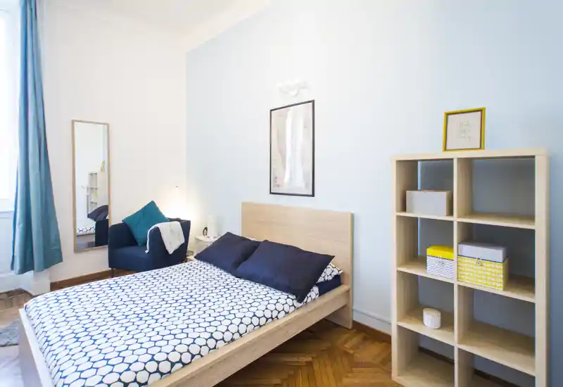 Camera da letto luminosa e moderna con letto in legno, biancheria a motivo e una libreria a cubi; ambiente arioso con luce naturale e arredo curato, perfetto per annunci.