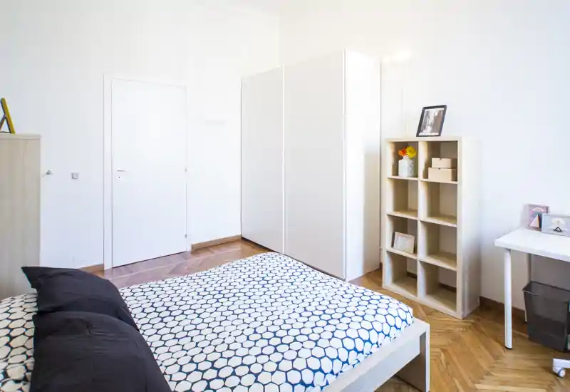Camera da letto luminosa e moderna con letto matrimoniale a fantasia, armadio bianco e scaffalatura; pavimento in parquet e scrivania essenziale. Perfetta per annunci che mostrano un'area notte ordinata e ben illuminata.