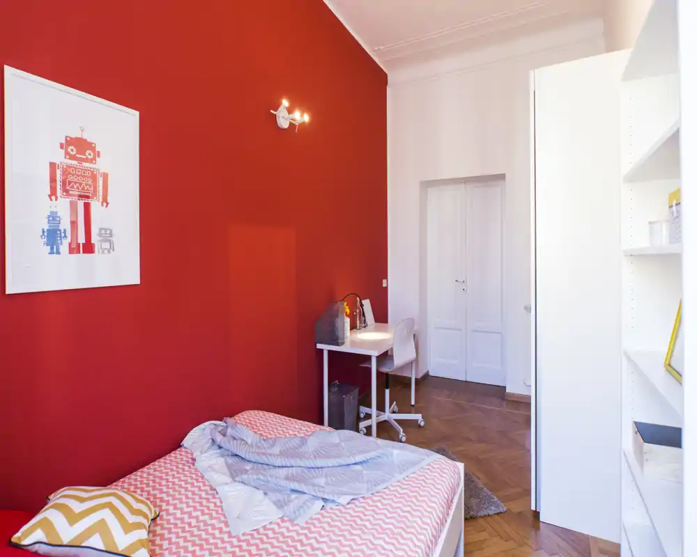 Chambre lumineuse et stylée avec un mur rouge vif, lit simple aux draps à motifs, petit bureau et étagères — idéale pour étudiant ou personne seule.