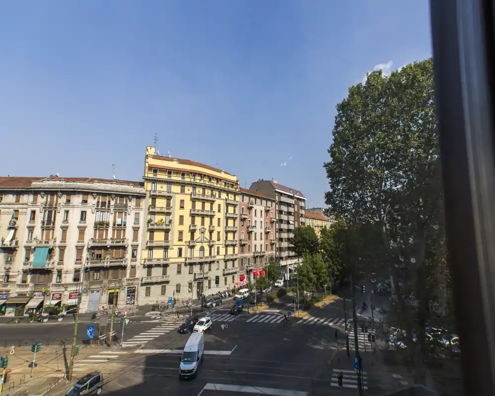 Vue extérieure sur la rue depuis une fenêtre montrant un carrefour ensoleillé, des immeubles résidentiels et de grands arbres.