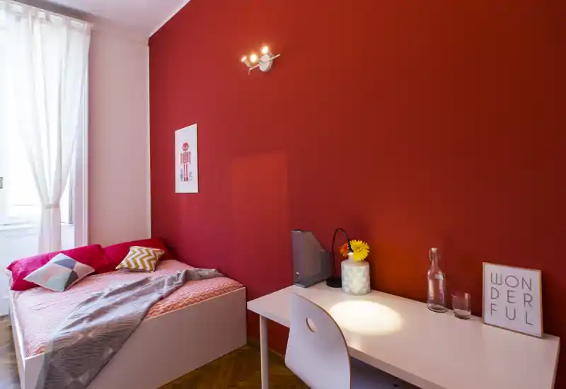 Camera da letto singola luminosa e accogliente con parete rossa a contrasto, letto con cuscini decorativi e una scrivania ordinata ideale per lo studio o il lavoro da remoto.
