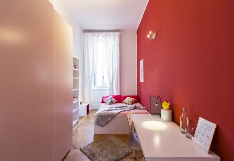 Camera singola luminosa e ben arredata con parete rossa, letto comodo, scrivania e luce naturale dalla finestra — perfetta per chi studia o lavora.