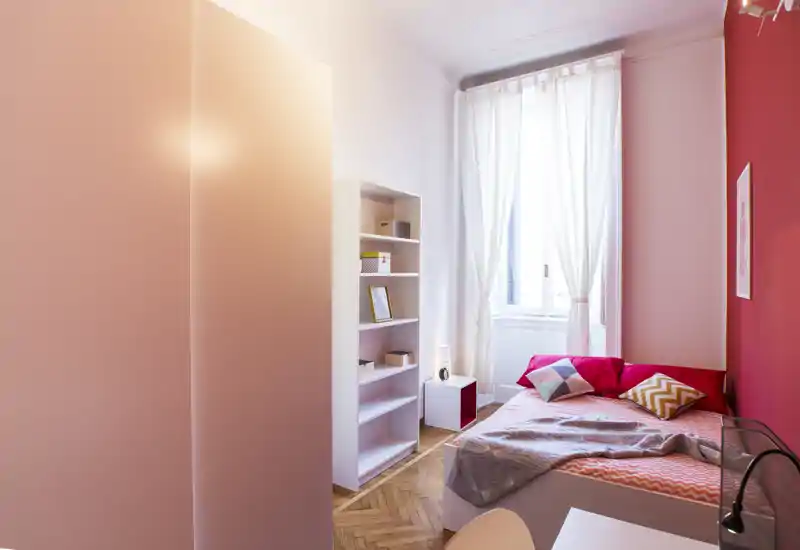 Camera da letto luminosa e di stile con letto singolo ben rifinito, cuscini decorativi e parete accentata rosa/rossa; finestra alta con tende leggere che diffondono luce naturale.