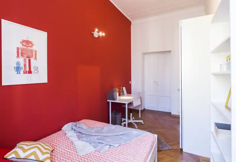Bright, stylish bedroom with a bold red accent wall, a neatly made single bed with patterned linens, a small desk and shelving — ideal for a student or single occupant.