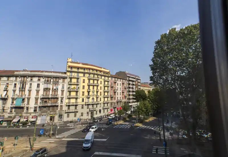 Street-facing outdoor view from a window showing a sunny intersection, multi-story residential buildings and mature trees.