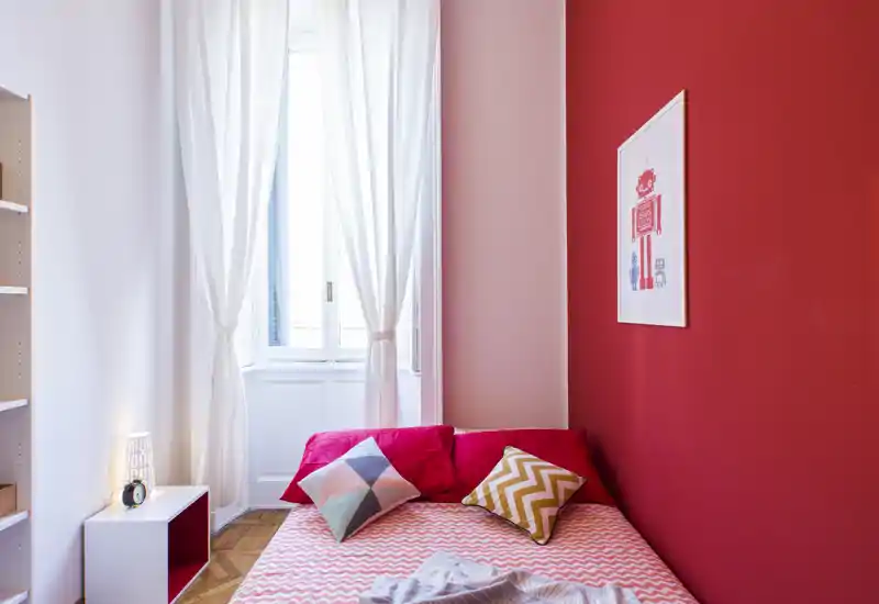 Camera da letto luminosa e compatta con letto matrimoniale, biancheria a fantasia, cuscini colorati e una grande finestra con tende leggere che illuminano la stanza di luce naturale.