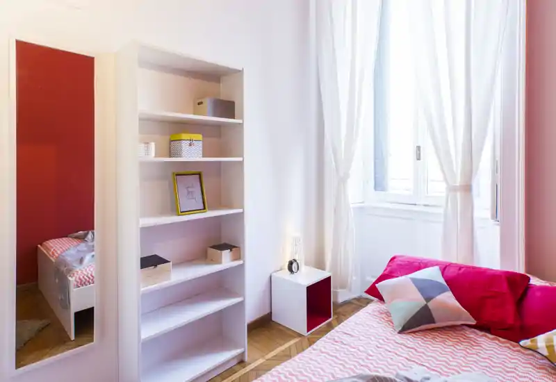 Camera da letto luminosa e ben arredata con copriletto a motivo, cuscini colorati e una grande finestra che illumina l'ambiente. Una libreria bianca, un comodino cubo e uno specchio a figura intera completano l'allestimento accogliente e contemporaneo.