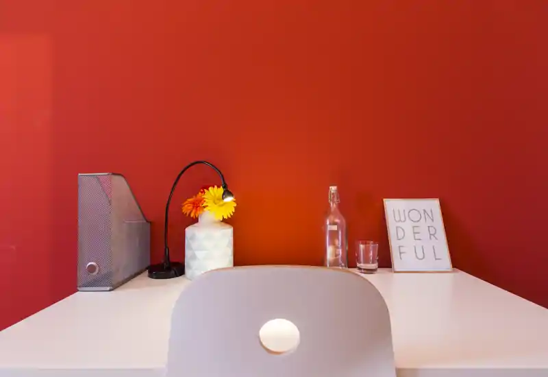 Compact workspace with a white desk against a warm red wall, featuring a small desk lamp, decorative vase with flowers, a water bottle and a framed print — clean, modern and inviting for work or study.