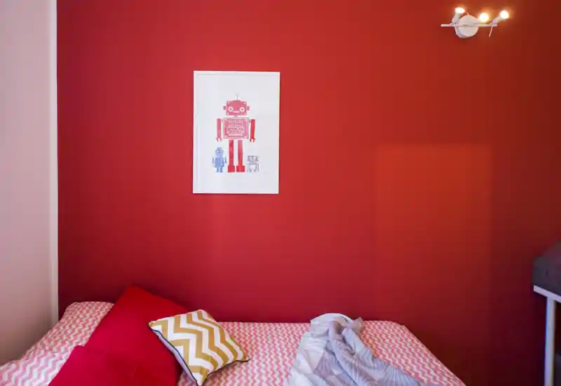 Bright, cozy bedroom corner with a patterned duvet, colorful cushions and a bold red accent wall featuring playful robot artwork.