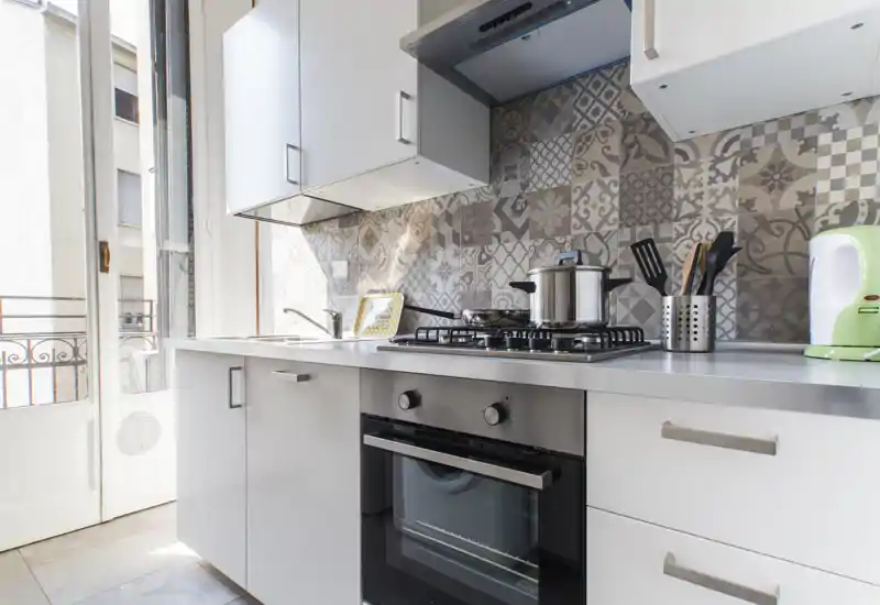 Bright modern kitchen with a built-in oven, gas hob and decorative patterned tile backsplash. Natural light enters from the balcony door, creating an airy, functional cooking space.