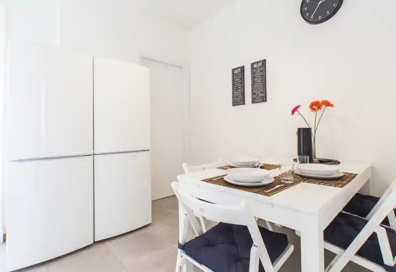 Compact, modern dining corner with a white table set for four, dark seat cushions and fresh flowers—bright, minimal and welcoming.