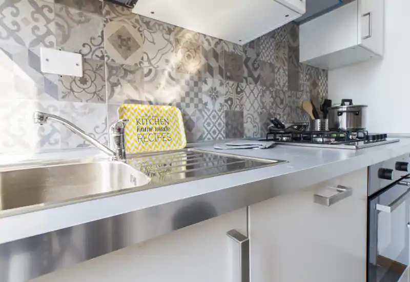 Modern compact kitchen with stainless-steel sink and gas hob, decorative patterned tile backsplash and clean white cabinetry — bright and neatly styled for rental listings.